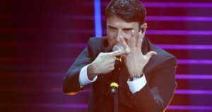 Una foto di scena dal palco del teatro Ariston durante Sanremo 2026: il vincitore, Sal Da Vinci, indica con lindice della mano destra l'anello nuziale che indossa sull'anulare della mano sinistra aperta davanti alla faccia, con un'espressione aggressiva sul volto.