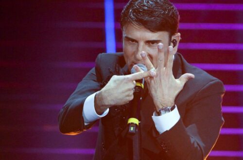 Una foto di scena dal palco del teatro Ariston durante Sanremo 2026: il vincitore, Sal Da Vinci, indica con lindice della mano destra l'anello nuziale che indossa sull'anulare della mano sinistra aperta davanti alla faccia, con un'espressione aggressiva sul volto.