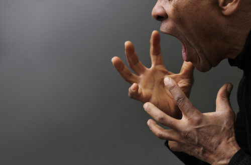 Una foto a colori, in formato orizzontale, che ritrae a destra un uomo di profilo, col volto rivolto verso sinistra, la bocca spalancata in un urlo di rabbia, le mani aperte con le dita ad artigliare l'aria poco sotto la faccia, davanti a uno sfondo che da grigio chiaro in alto a sinistra diventa gradualmente grigio scuro in basso a destra.