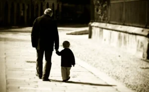Una foto in bianco e nero, orizzontale, che ritrae di spalle un vecchio signore alto che cammina tenendo per mano un bambino molto piccolo in mezzo a una strada sgombra con pavè e pietre bianche.