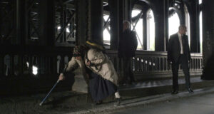 Un fotogramma dal film “Holy Motors”, di Leos Carax. Sul marciapiedi di una strada che passa sotto un grande ponte di metallo incede una mendicante infagottata in vestiti logori e pesanti, con un foulard sulla testa e uno zainetto sulla schiena tutta incurvata in avanti, puntellandosi con una stampella stretta nella mano destra protesa in avanti. La mendicante è seguita a pochi passi da due guardie del corpo in abiti eleganti.