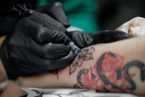 Una foto a colori, in formato orizzontale, che ritrae da vicino la mano destra di un tatuatore, avvolta in un guanto di gomma nera, mentre traccia un tatuaggio sull'avambraccio di qualcuno.