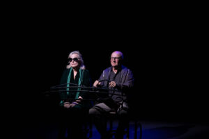 Foto di scena dallo spettacolo teatrale "Terzo Tempo". Al centro del palco, davanti a uno sfondo completamente buio, ci sono Lucia Vasini e Paolo Hendel che fingono di andare in auto (alla guida c'è Hendel). La foto è di Asia Ludovica Serpe.