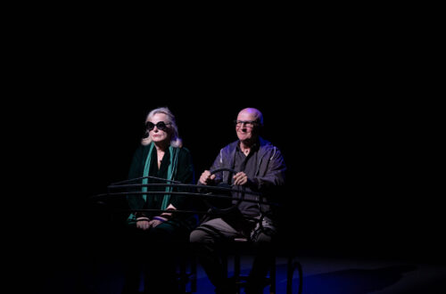 Foto di scena dallo spettacolo teatrale "Terzo Tempo". Al centro del palco, davanti a uno sfondo completamente buio, ci sono Lucia Vasini e Paolo Hendel che fingono di andare in auto (alla guida c'è Hendel). La foto è di Asia Ludovica Serpe.