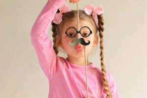 Una foto che ritrae al centro una bambina vestita di rosa e con lunghe trecce che le scendono ai lati del volto mentre per gioco finge di essere un uomo con un paio di occhiali neri rotondi e un paio di baffi altrettanto neri e posticci, sotto i quali manda un bacino verso l'obiettivo.