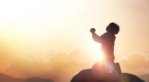 Una foto che ritrae un bambino in ginocchio in cima a una roccia, davanti a un tramonto, mentre prega con la testa leggermente inclinata indietro e le mani giunte davanti a sé un po' sollevate verso il cielo.