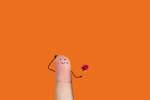 Una foto che ritrae al centro l’estremità di un dito umano, un indice, teso verso l’alto davanti a uno sfondo arancione. Sulla falange del dito sono disegnati, con tratto molto infantile, un paio di occhi, una bocca sorridente e due gote rosse ai suoi lati. Poco al di sotto di questo viso, dai lati del dito escono due braccia: la sinistra porta la mano al lato sinistro della “testa”, con un gesto che appare un po’ vezzoso; la destra regge davanti al “busto” una rosa di colore rosa. Nell’insieme il dito sembra prepararsi a un incontro galante.