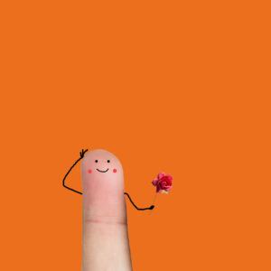Una foto che ritrae al centro l’estremità di un dito umano, un indice, teso verso l’alto davanti a uno sfondo arancione. Sulla falange del dito sono disegnati, con tratto molto infantile, un paio di occhi, una bocca sorridente e due gote rosse ai suoi lati. Poco al di sotto di questo viso, dai lati del dito escono due braccia: la sinistra porta la mano al lato sinistro della “testa”, con un gesto che appare un po’ vezzoso; la destra regge davanti al “busto” una rosa di colore rosa. Nell’insieme il dito sembra prepararsi a un incontro galante.