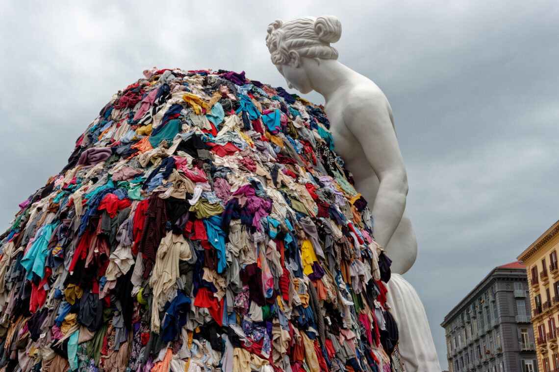 Napoli, Italia - 30 settembre 2023: La monumentale installazione artistica “Venere degli stracci” dell’artista contemporaneo Michelangelo Pistoletto è esposta all’aperto in città. Una classica scultura bianca raffigurante la dea romana si affaccia su un’enorme pila di indumenti colorati scartati. Questo iconico capolavoro dell’Arte Povera contrappone visivamente l’antica bellezza classica al consumismo e agli sprechi della moda fast fashion moderna, offrendo una potente riflessione culturale e sociale ai turisti di tutto il mondo e agli amanti dell’arte.