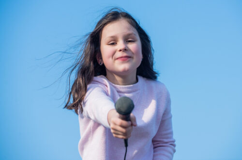 Una foto che ritrae al centro il mezzobusto di una ragazzina sorridente che protende davanti a sé un microfono, come a voler intervistare chi ha davanti.