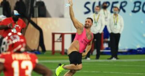 Una foto a colori che ritrae al centro un uomo mezzo nudo che corre in mezzo a un campo da football americano, sorridendo verso l'obiettivo, inseguito da alcuni uomini della security.