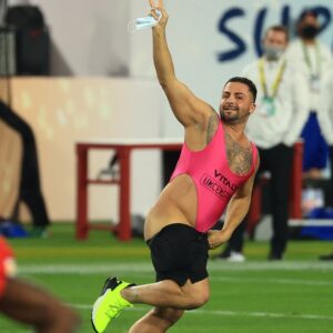 Una foto a colori che ritrae al centro un uomo mezzo nudo che corre in mezzo a un campo da football americano, sorridendo verso l'obiettivo, inseguito da alcuni uomini della security.