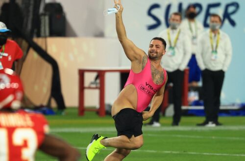 Una foto a colori che ritrae al centro un uomo mezzo nudo che corre in mezzo a un campo da football americano, sorridendo verso l'obiettivo, inseguito da alcuni uomini della security.