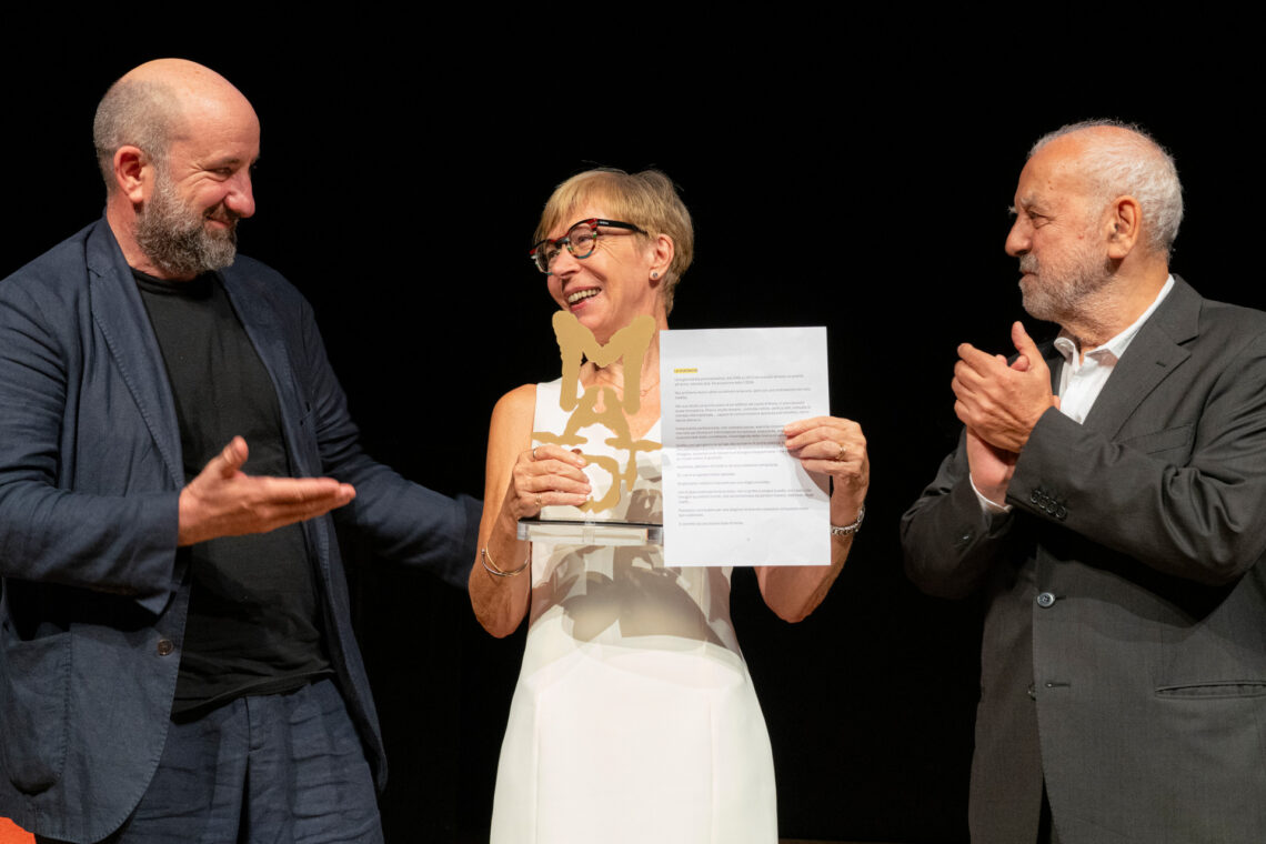 Una foto a colori, in formato orizzontale, che ritrae, da sinistra a destra, Antonio Albanese, Milena Gabanelli e Giampietro Savuto sul palco del Teatro Gerolamo, bene illuminati davanti a uno sfondo completamente nero. Antonio Albanese è di profilo e guarda sorridendo Milena Gabanelli, alla sua sinistra (a destra nell'inquadratura), la quale a sua volta gli sorride reggendo con la mano destra una piccola scultura, il primo appena ricevuto dalle mani di Albanese, e con la mano sinistra l'attestato relativo. Alla sinistra della Gabanelli (ulteriormente a destra nell'inquadratura), Giampietro Savuto la guarda con un'espressione contenta, forse un po' commossa, mentre applaude con le mani all'altezza del petto.