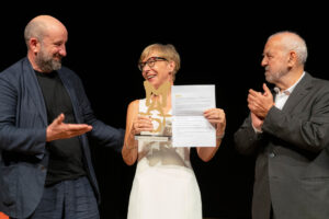 Una foto a colori, in formato orizzontale, che ritrae, da sinistra a destra, Antonio Albanese, Milena Gabanelli e Giampietro Savuto sul palco del Teatro Gerolamo, bene illuminati davanti a uno sfondo completamente nero. Antonio Albanese è di profilo e guarda sorridendo Milena Gabanelli, alla sua sinistra (a destra nell'inquadratura), la quale a sua volta gli sorride reggendo con la mano destra una piccola scultura, il primo appena ricevuto dalle mani di Albanese, e con la mano sinistra l'attestato relativo. Alla sinistra della Gabanelli (ulteriormente a destra nell'inquadratura), Giampietro Savuto la guarda con un'espressione contenta, forse un po' commossa, mentre applaude con le mani all'altezza del petto.