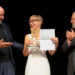 Una foto a colori, in formato orizzontale, che ritrae, da sinistra a destra, Antonio Albanese, Milena Gabanelli e Giampietro Savuto sul palco del Teatro Gerolamo, bene illuminati davanti a uno sfondo completamente nero. Antonio Albanese è di profilo e guarda sorridendo Milena Gabanelli, alla sua sinistra (a destra nell'inquadratura), la quale a sua volta gli sorride reggendo con la mano destra una piccola scultura, il primo appena ricevuto dalle mani di Albanese, e con la mano sinistra l'attestato relativo. Alla sinistra della Gabanelli (ulteriormente a destra nell'inquadratura), Giampietro Savuto la guarda con un'espressione contenta, forse un po' commossa, mentre applaude con le mani all'altezza del petto.