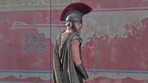Un fotogramma dal film “Odissea”, di Cristopher Nolan, in cui al centro, davanti a un muro dipinto di rosso tenue, un po’ scrostato, c’è Ulisse voltato, quasi di spalle, con la testa girata verso la sua destra e quindi di profilo, coperta da un pesante elmo metallico con un’alta cresta a spazzola di pelo rosso, e un’espressione molto seria e un po’ malinconica sul volto.