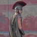 Un fotogramma dal film “Odissea”, di Cristopher Nolan, in cui al centro, davanti a un muro dipinto di rosso tenue, un po’ scrostato, c’è Ulisse voltato, quasi di spalle, con la testa girata verso la sua destra e quindi di profilo, coperta da un pesante elmo metallico con un’alta cresta a spazzola di pelo rosso, e un’espressione molto seria e un po’ malinconica sul volto.